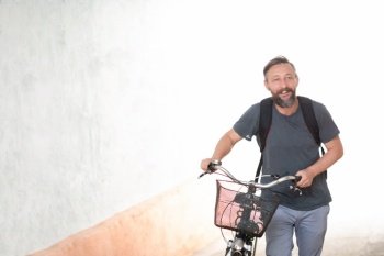 A young smiling stylish bearded hipster man with a backpack pushing a retro bicycle while walking on street