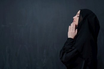 portrait of beautiful muslim woman in fashionable dress with hijab making traditional prayer to God keeps hands in praying gesture isolated on black chalkboard background representing modern islam fashion and ramadan kareem concept