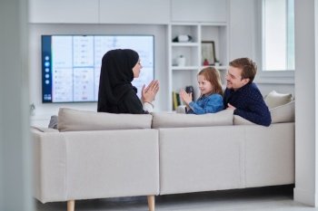 Happy Muslim family spending time together in modern home. Parents playing with daughter on the sofa during Ramadan. Traditional fashionable arabic clothes.