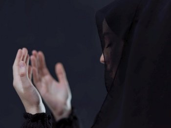 portrait of beautiful muslim woman in fashionable dress with hijab making traditional prayer to God, keeps hands in praying gesture isolated on black chalkboard background representing modern islam fashion and ramadan kareem concept