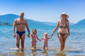 portrait of a young happy couple with cute little daughters having fun while standing in the shallow water of the sea during Summer vacation  Healthy family holiday concept