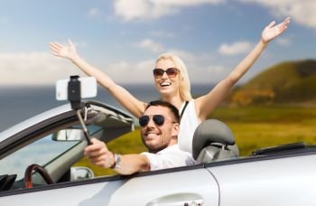 road trip, technology and travel concept - happy couple driving in convertible car and taking picture by smartphone on selfie stick. happy couple in car taking selfie by smartphone