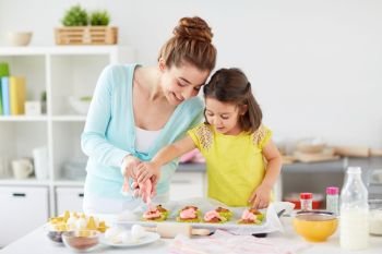 family, cooking and people concept - mother and little daughter making and decorating cupcakes with cream frosting at home kitchen. mother and daughter cooking cupcakes at home