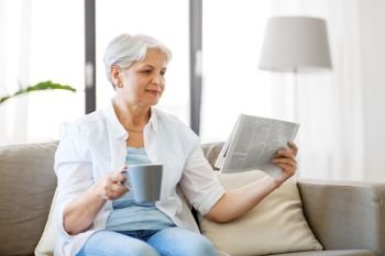 old age, leisure and people concept - happy senior woman drinking coffee or tea and reading newspaper at home. senior woman with coffee reading newspaper at home