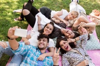 friendship, leisure and technology concept - group of happy smiling friends taking selfie by smartphone chilling on picnic blanket at summer park. friends taking selfie by smartphone at picnic