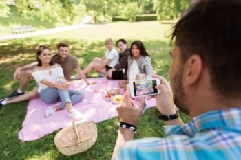 friendship, leisure and technology concept - man taking picture of his friends by smartphone on picnic at summer park. man photographing friends by smartphone at picnic