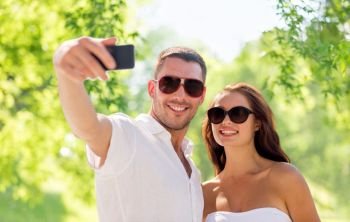 love, summer and technology concept - smiling couple in sunglasses making selfie by smartphone over green natural background. couple making selfie over natural background
