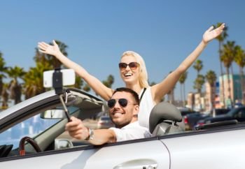 road trip, technology and travel concept - happy couple driving in convertible car and taking picture by smartphone on selfie stick over venice beach background in california. happy couple in car taking selfie by smartphone