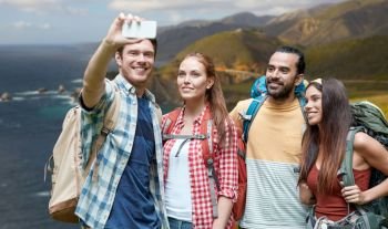 technology, travel, tourism, hike and people concept - group of smiling friends with backpacks taking selfie by smartphone over bixby creek bridge on big sur coast of california background. friends with backpack taking selfie by smartphone