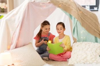 childhood, friendship and hygge concept - happy little girls reading book with torch light in kids tent or teepee at home. little girls reading book in kids tent at home