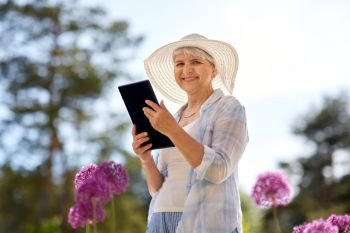 gardening, technology and people concept - happy senior woman with tablet pc computer and flowers at summer garden. senior woman with tablet pc and flowers at garden