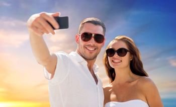love, summer and technology concept - smiling couple in sunglasses making selfie by smartphone over sky sunset background. couple making selfie by smartphone over sunset sky