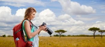 travel, tourism and photography concept - happy young woman with backpack and camera photographing over african savannah background. woman with backpack and camera over savannah