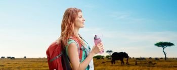 adventure, travel, tourism, hike and people concept - smiling young woman with backpack and bottle of water over animals in african savannah background. happy woman with backpack over african savannah