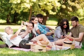friendship, leisure, technology and people concept - group of friends with smartphones chilling on picnic blanket and drinking non alcoholic beer at summer park. friends with smartphones on picnic at summer park
