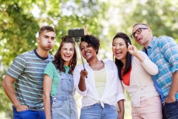 people, friendship and international concept - happy smiling young woman and group of happy friends taking picture by selfie stick in park. international friends taking selfie in park