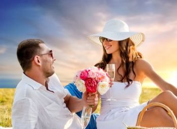 love, dating, people and celebration concept - smiling couple with flowers drinking champagne at picnic on meadow over sunset sky background. couple with flowers drinking champagne at picnic