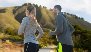fitness, sport and technology concept - happy couple running and listening to music in earphones over big sur hills background in california. couple with earphones running over big sur