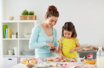 family, cooking and people concept - happy mother and little daughter making and decorating cupcakes with chocolate sprinkles at home kitchen. happy mother and daughter cooking cupcakes at home