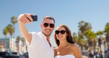 travel, tourism and technology concept - smiling couple in sunglasses making selfie by smartphone over venice beach background in california. couple in shades making selfie over venice beach
