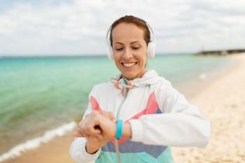 sport, technology and healthy lifestyle concept - smiling woman with headphones and fitness tracker exercising on beach. woman with headphones and fitness tracker on beach