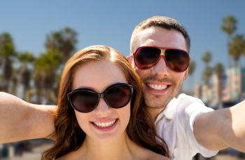 travel, tourism and people concept - smiling couple wearing sunglasses making selfie over venice beach background in california. couple in shades making selfie over venice beach