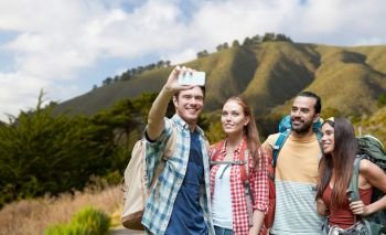 technology, travel, tourism, hike and people concept - group of smiling friends with backpacks taking selfie by smartphone over big sur hills of california background. friends with backpack taking selfie by smartphone