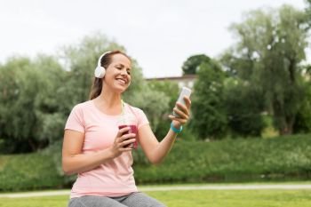 fitness, technology and healthy lifestyle concept - woman with smoothie drink or shake in plastic cup, headphones and smartphone listening to music after exercising in park. woman with smartphone and shake listening to music