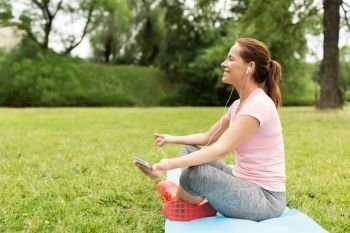 fitness, sport and technology concept - woman with smartphone and earphones meditating on yoga mat at park. woman meditating on yoga mat at park