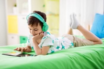 childhood, technology and people concept - smiling boy with tablet pc computer and headphones lying on bed at home. smiling boy with tablet pc and headphones at home