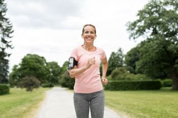 fitness, sport and healthy lifestyle concept - smiling woman with earphones wearing armband with smartphone and jogging at park and listening to music. woman with earphones and armband jogging at park