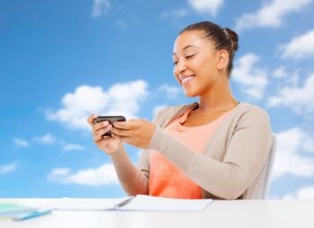 technology, education and people concept - smiling african student girl with smartphone and notebooks over blue sky background. african student girl with smartphone and notebooks