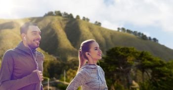 fitness, sport and technology concept - happy couple running and listening to music in earphones over big sur hills background in california. couple with earphones running over big sur hills