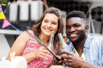 leisure, technology and people concept - happy mixed race couple with drinks taking picture by selfie stick at food truck. mixed race couple taking picture by selfie stick