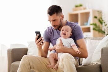 family, parenthood and technology concept - father with crying little baby and smartphone at home. father with crying baby and smartphone at home