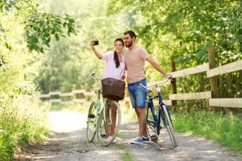 people, technology and lifestyle concept - happy couple with bicycles taking selfie by smartphone at summer park. couple with bicycles taking selfie by smartphone