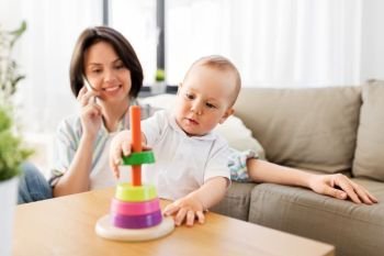 family, child and motherhood concept - happy baby boy playing developmental toy and mother calling on smartphone at home. baby boy playing toy and mother calling on phone