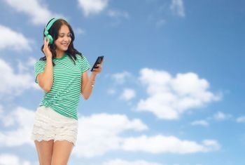 music, technology and people concept - happy teenage girl in headphones with smartphone over blue sky and clouds background. teenage girl in phones with smartphone over sky
