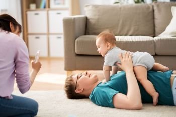 family, technology and people concept - happy mother and father taking picture or recording video of baby boy with smartphone at home. mother picturing father with baby by smartphone