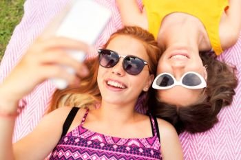 summer fashion, eyewear and leisure concept - smiling teenage girls in sunglasses lying on picnic blanket taking selfie. teenage girls in sunglasses taking selfie
