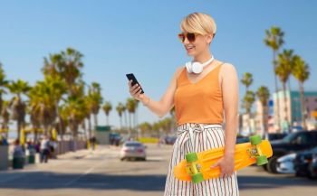 sport, leisure and skateboarding concept - smiling teenage girl in sunglasses with short modern cruiser skateboard and smartphone over venice beach background in california. teenage girl with skateboard and smartphone