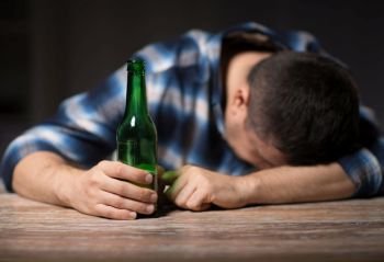 alcoholism, alcohol addiction and people concept - male alcoholic with beer bottles lying or sleeping on table at night. drunk man with beer bottles on table at night