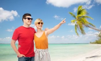 travel, tourism and summer holidays concept - smiling couple in sunglasses hugging over exotic tropical beach with palm trees background and pointing finger to something. smiling couple in sunglasses hugging