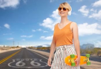 sport, leisure and skateboarding concept - smiling teenage girl in sunglasses and headphones with short modern cruiser skateboard over route 66 background. smiling teenage girl with skateboard over