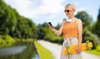 sport, leisure and skateboarding concept - smiling teenage girl in sunglasses with short modern cruiser skateboard and smartphone over summer park background. teenage girl with skateboard and smartphone
