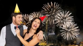 birthday, celebration and holidays concept - happy couple with party blowers and caps having fun over firework lights at night city background. happy couple with party blowers having fun