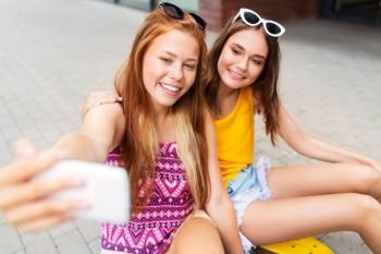 friendship, leisure and technology concept - happy teenage girls or friends with skateboards on city street taking selfie by smartphone in summer. teenage girls taking selfie on city street