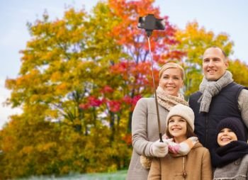 family, technology and people concept - happy mother, father, daughter and son taking picture by smartphone on selfie stick over autumn park background. family taking selfie over autumn park background