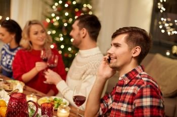 holidays, communication and celebration concept - happy young man calling on smartphone and having christmas dinner with friends at home. man calling on smartphone at christmas dinner