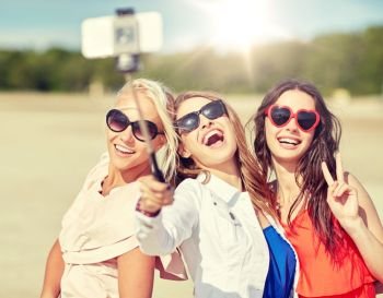 summer vacation, holidays, travel, technology and people concept- group of smiling young women taking picture with smartphone on selfie stick on beach. group of smiling women taking selfie on beach